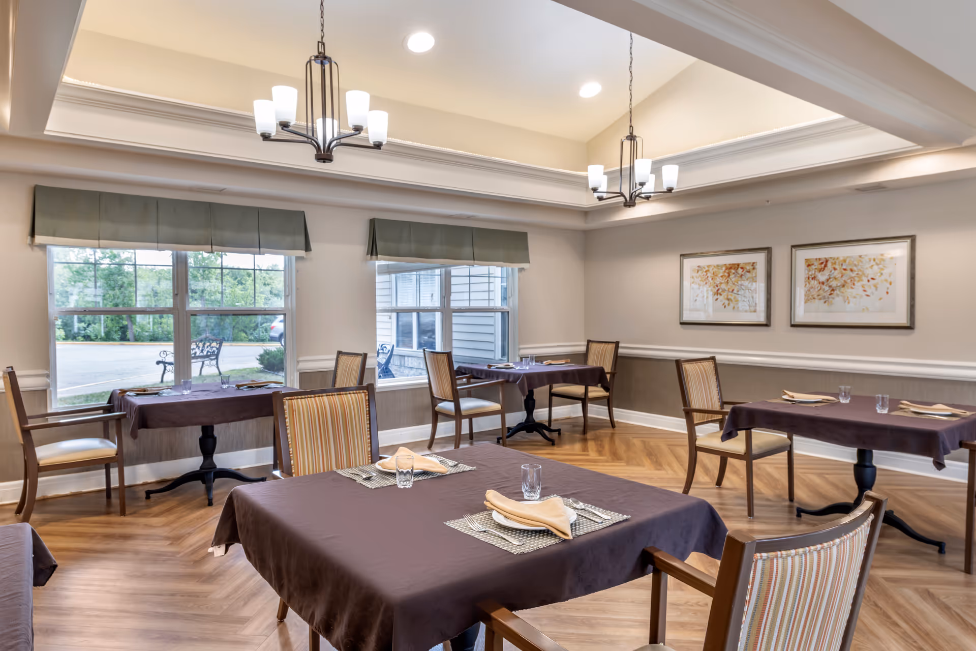 A dining room with several tables covered in dark tablecloths, each set with placemats, napkins, and glasses. The room has large windows with green valances, wooden chairs with striped cushions, two framed artworks on the wall, and two ceiling light fixtures with multiple bulbs. The floor is wood with a herringbone pattern.