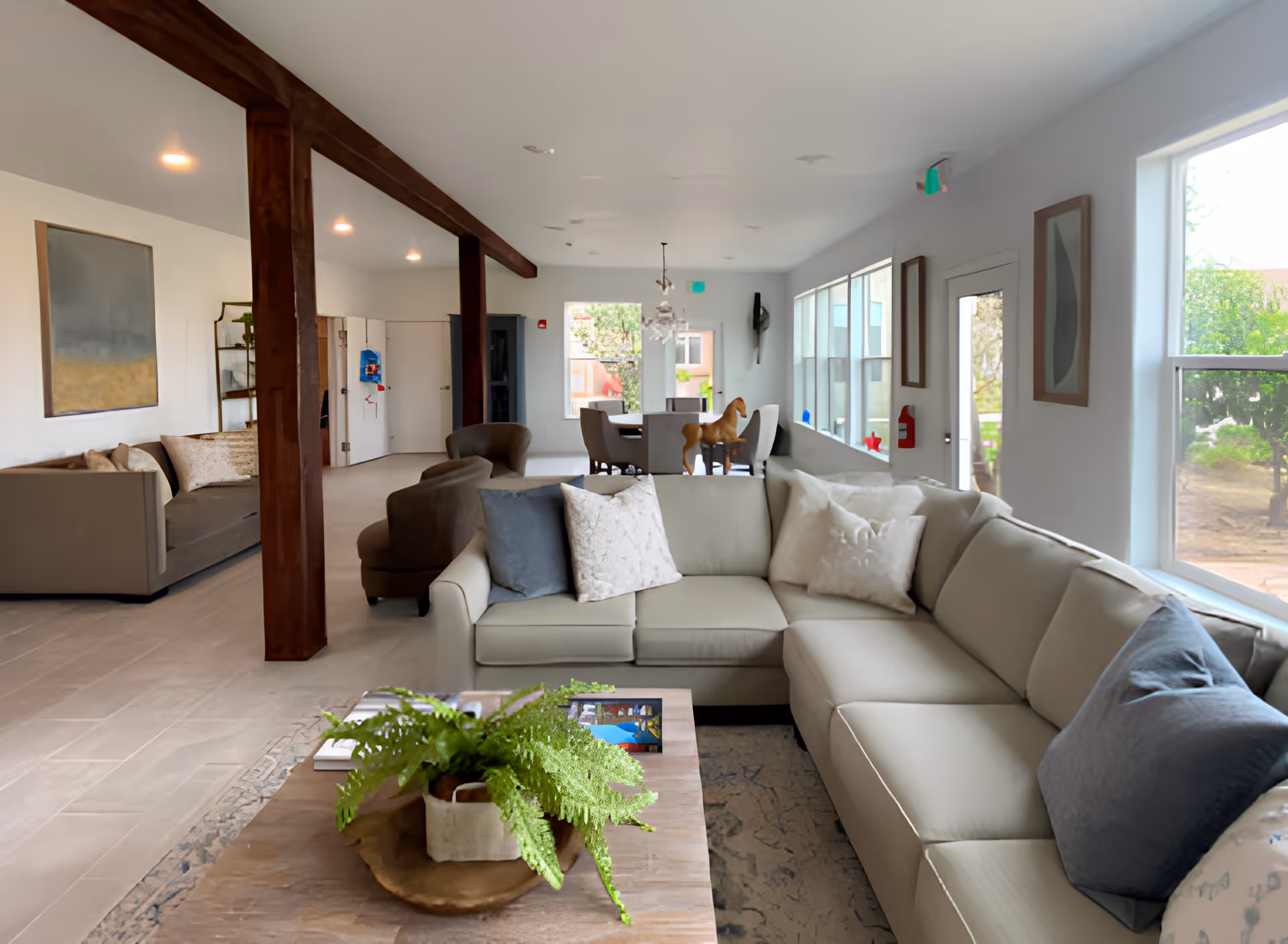 A spacious and well-lit living room area in a senior living facility with a large beige sectional sofa adorned with various pillows, a wooden coffee table with a green potted plant, and additional seating including armchairs and another sofa. The room features large windows letting in natural light, wooden beams on the ceiling, framed artwork on the walls, and a dining area with chairs and a wooden rocking horse in the background.