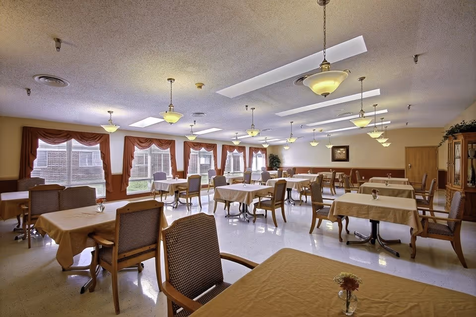 A spacious dining room with multiple tables covered in beige tablecloths, each surrounded by wooden chairs with cushioned seats. The room has large windows with red curtains allowing natural light to fill the space. Several hanging ceiling lights and skylights provide additional illumination. A wooden cabinet with decorative items is visible on the right side of the room.