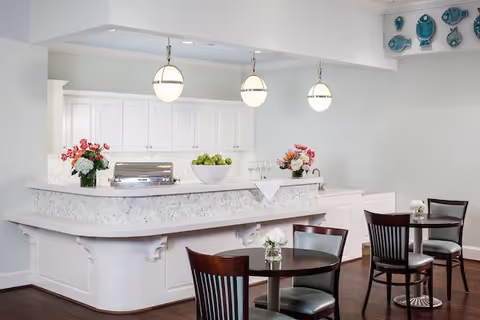 A bright and clean dining area with a white serving counter decorated with flowers and a bowl of green apples. Three round pendant lights hang above the counter. In front of the counter, there are small round tables with dark wooden chairs, each table adorned with a small flower arrangement. The walls are light-colored with decorative plates displayed on the upper right corner.