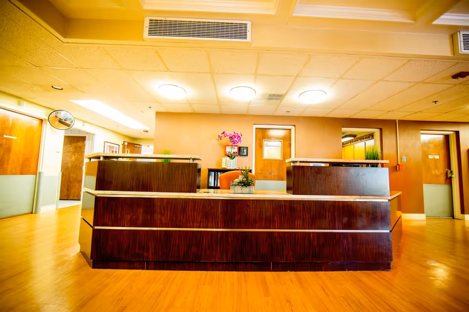 Reception desk area inside a senior living facility with a wooden counter, a small plant, and a purple orchid on the desk. The room has wooden floors, beige walls, ceiling lights, and several doors in the background.