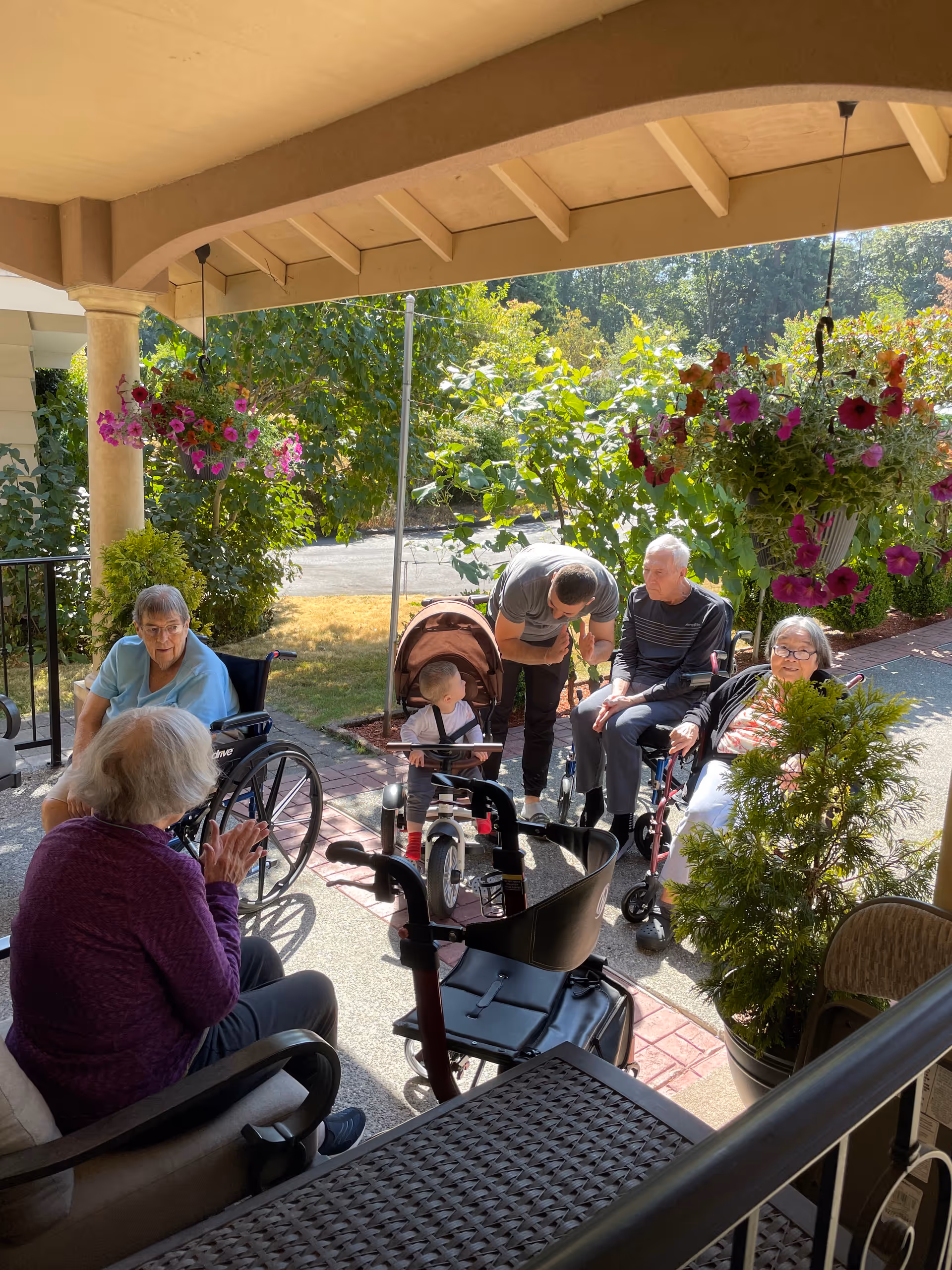 A group of elderly people and a young child are gathered outside on a covered patio area. Two elderly women are seated in wheelchairs, another elderly woman is sitting on a chair, and a man is bending down to interact with the child who is standing near a stroller. The patio is decorated with hanging flower baskets and potted plants, with greenery and trees visible in the background.