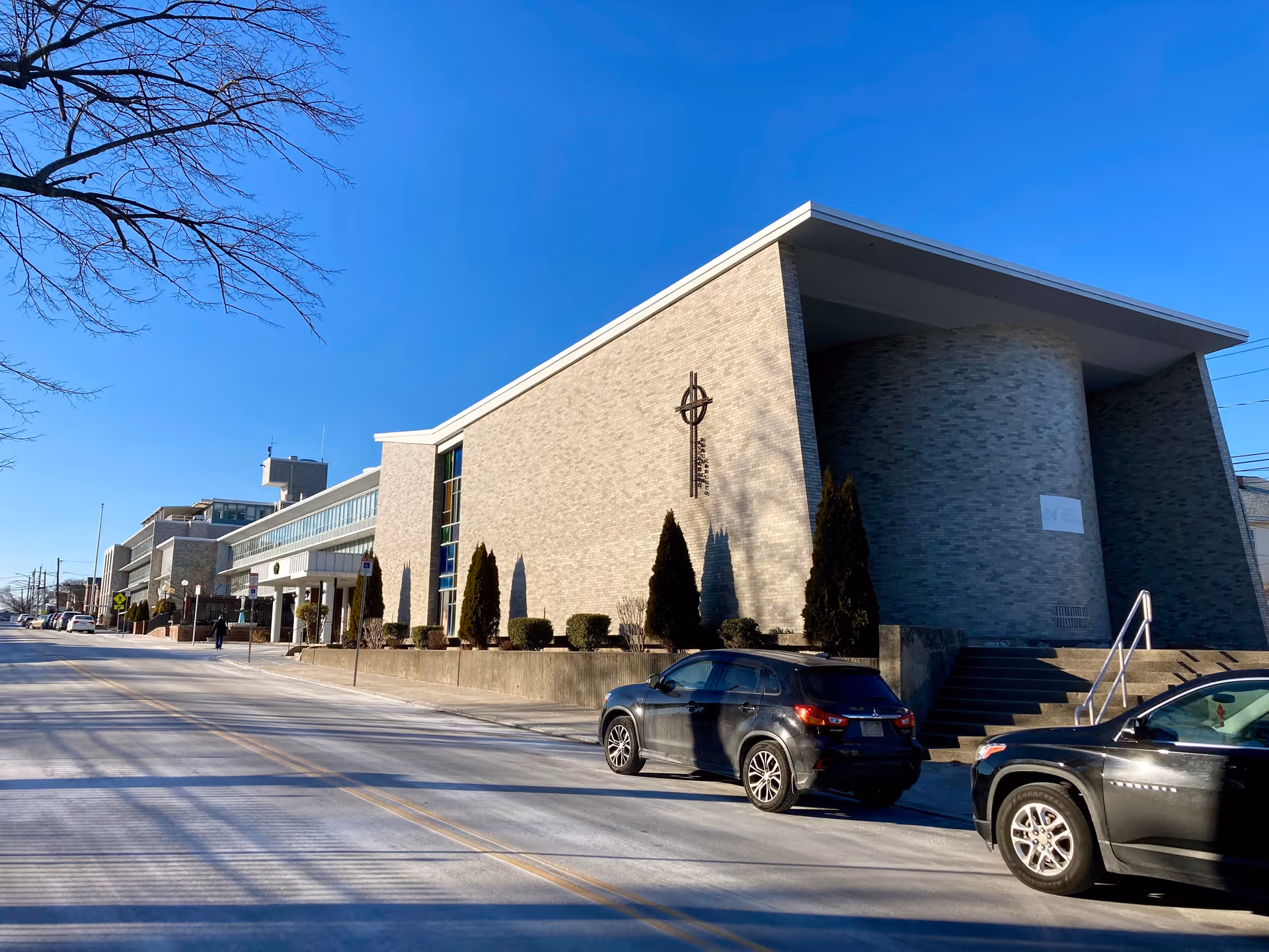 Exterior view of a large brick building with a modern architectural design, featuring a curved wall section and a cross symbol mounted on the facade. Two black cars are parked on the street in front of the building, and a clear blue sky is visible above.