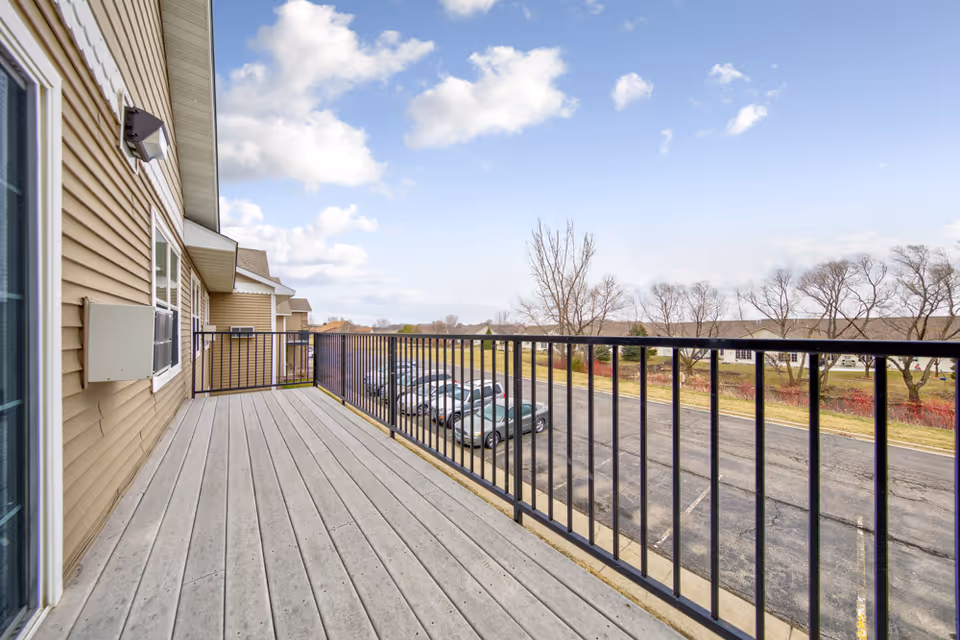 View from a second-floor balcony with wooden flooring and black metal railing overlooking a parking lot with several cars and a grassy area with trees under a partly cloudy sky.