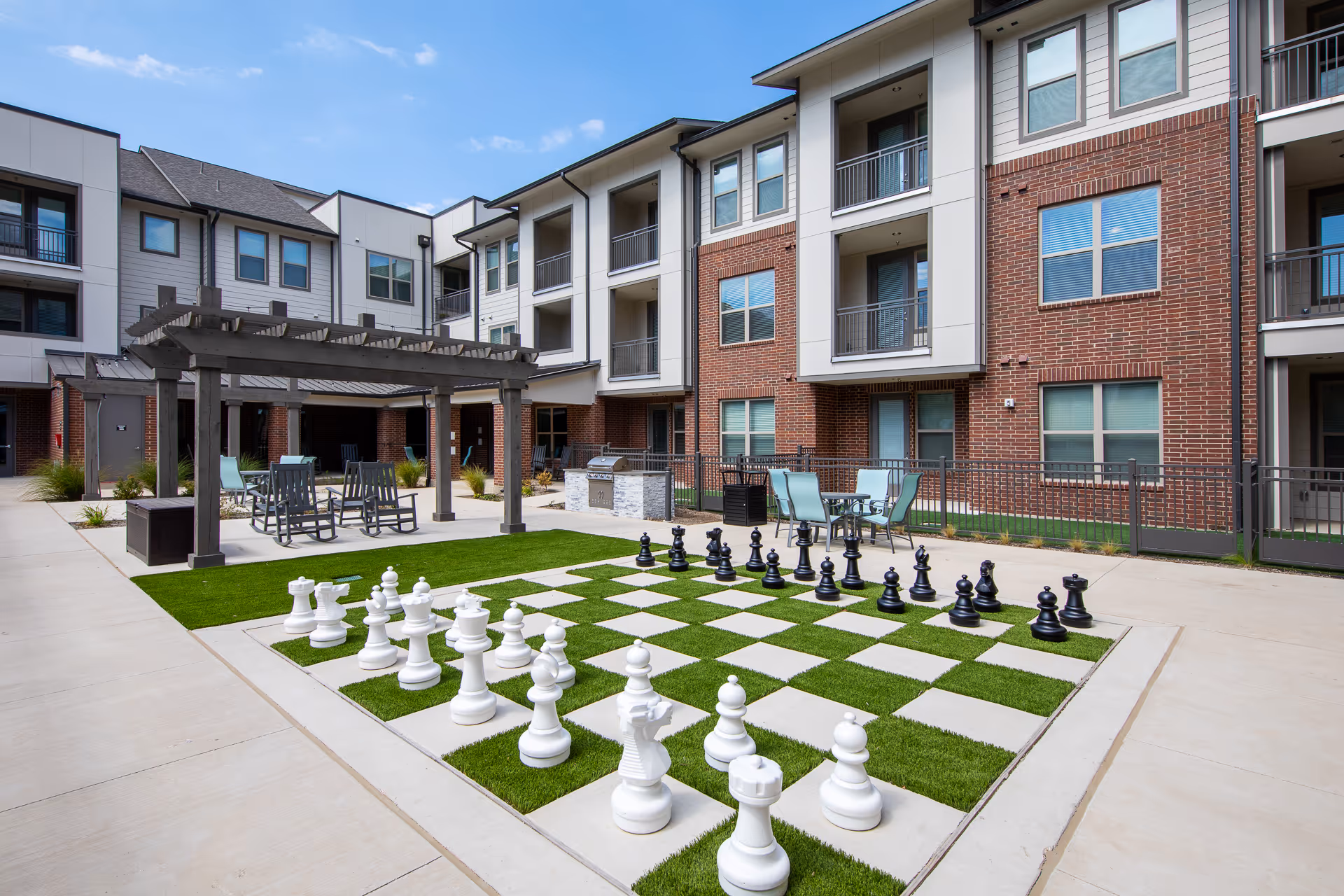 Outdoor courtyard with a large grass-and-tile giant chessboard, pergola and seating in front of a three-story apartment building.