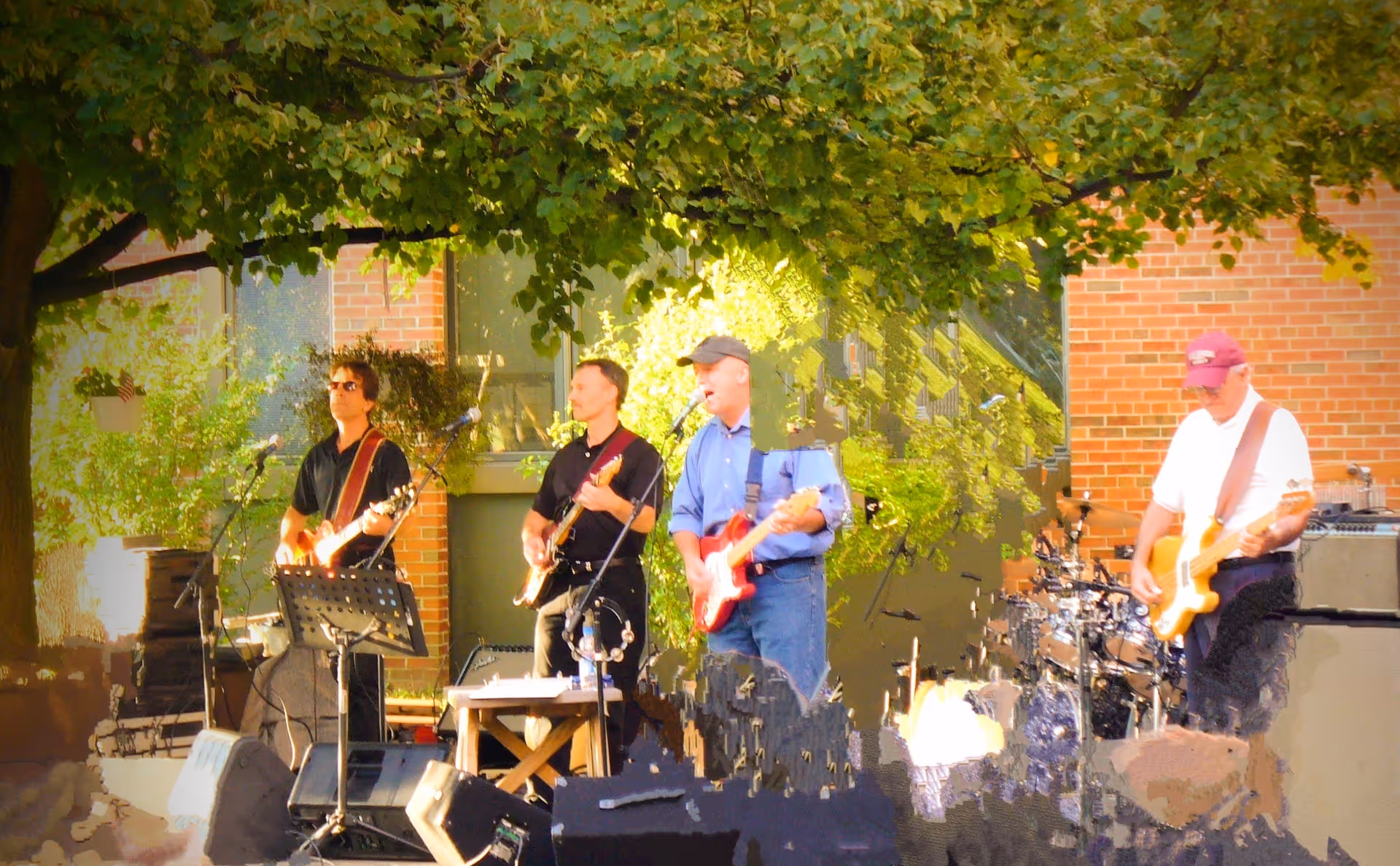 A small band of musicians playing guitars and drums outdoors beneath a tree in front of a brick building.