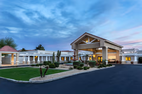 Exterior view of The Terraces of Phoenix senior living facility at dusk, featuring a covered entrance with columns, landscaped garden with shrubs and cacti, and a paved driveway under a partly cloudy sky.