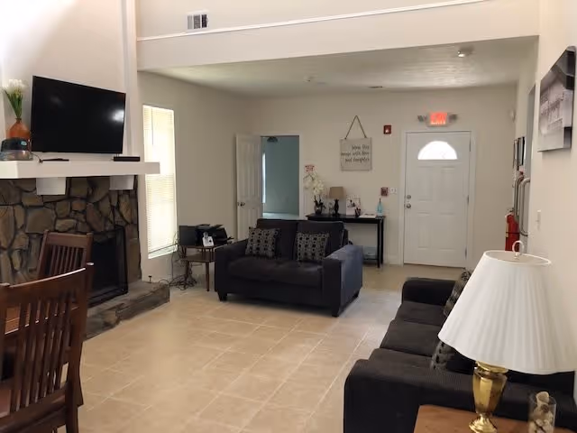 Interior view of a living room area with two dark gray sofas, a stone fireplace with a mounted flat-screen TV above it, a wooden dining table with chairs, a small table with a lamp, and a white door in the background. The room has beige tiled flooring and light-colored walls.