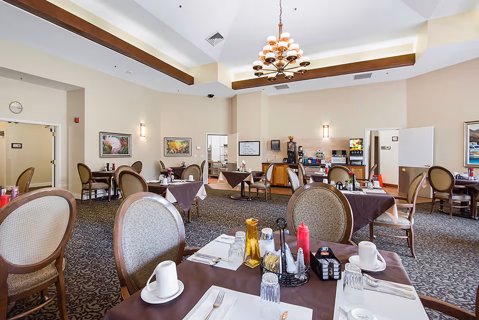 A spacious dining room with multiple tables covered in brown tablecloths and set with cups, glasses, utensils, and condiments. The room features upholstered chairs, a patterned carpet, a chandelier hanging from the ceiling, and a beverage station with coffee and soda machines against the far wall. There are framed paintings on the walls and doors leading to other rooms.