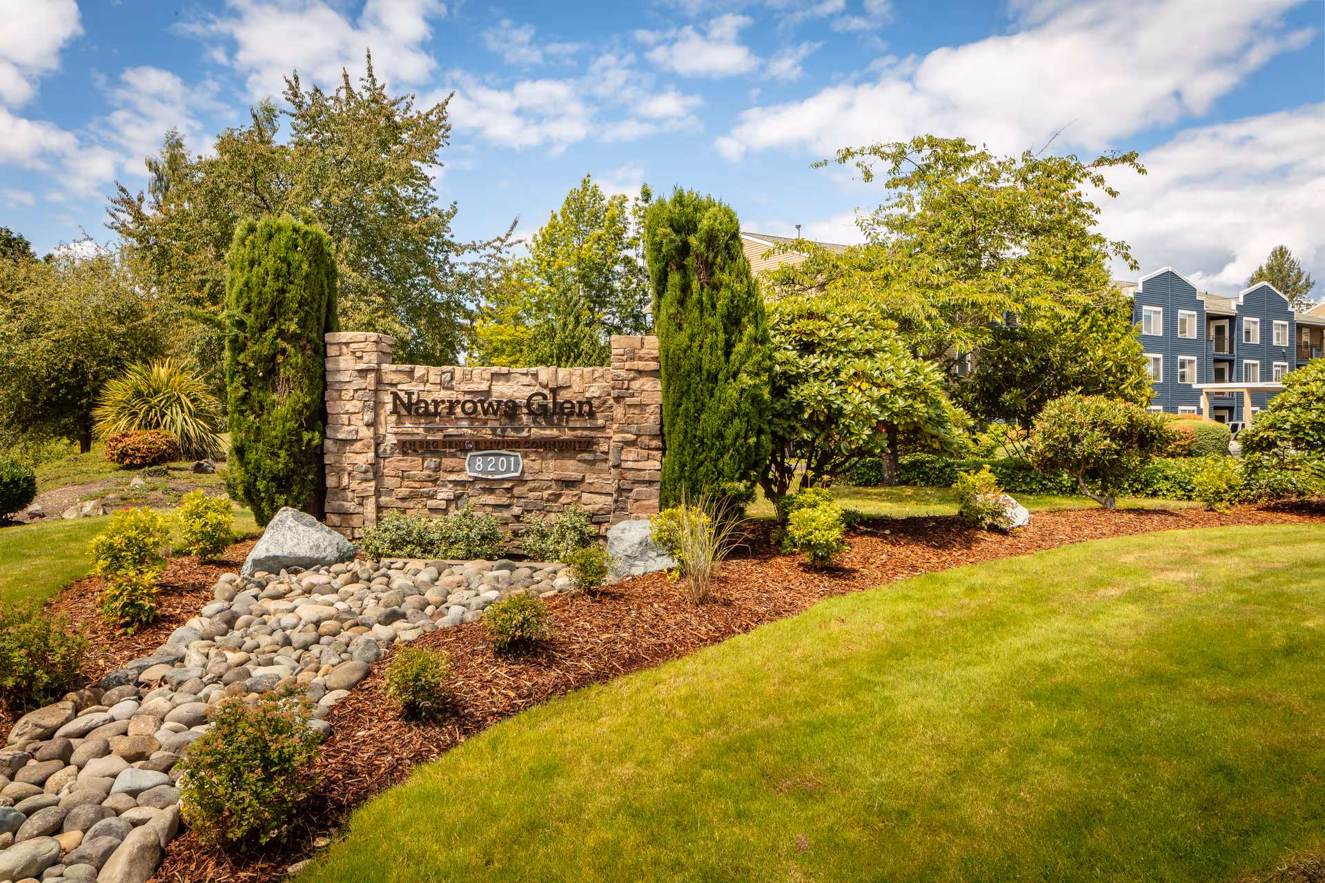 Stone sign for Narrows Glen senior living community surrounded by landscaped greenery, bushes, and trees with a blue multi-story building in the background under a partly cloudy sky.