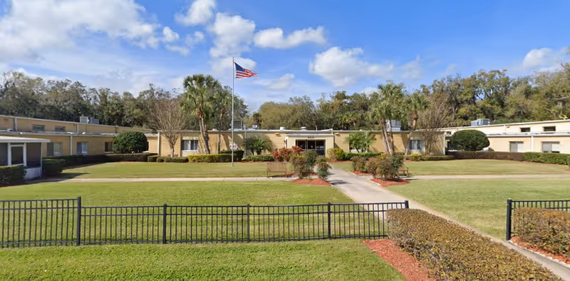 Front view of a single-story senior living facility with a flagpole, benches, and landscaped lawn.