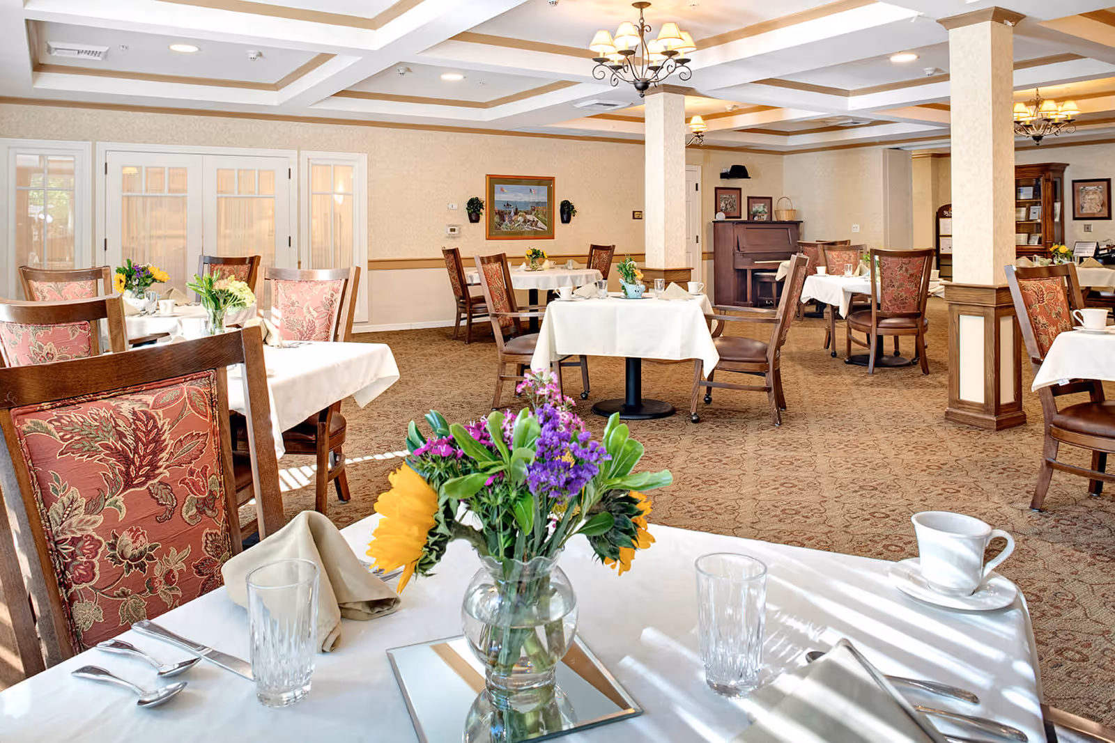 A bright and elegant dining room with tables covered in white tablecloths, each set with glassware, napkins, and silverware. Floral centerpieces decorate the tables. The room features patterned carpet, wooden chairs with floral upholstery, coffered ceiling with chandeliers, and framed artwork on the walls.