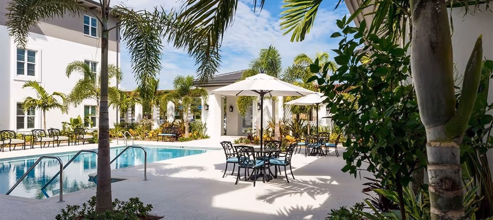 Outdoor pool and patio area with umbrella-shaded tables, lounge chairs, palm trees, and a neighboring building under a blue sky.
