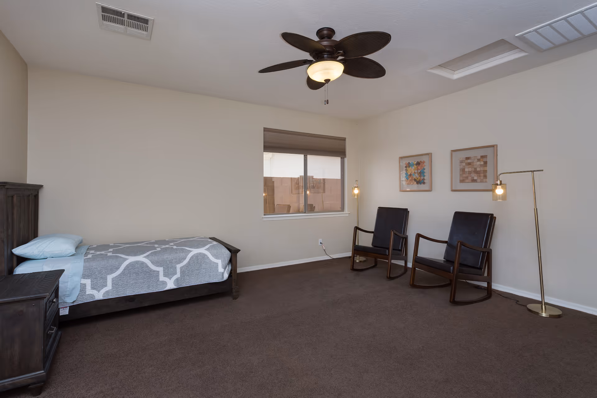 A simple bedroom with a single bed covered with a gray and white patterned blanket and a light blue pillow. Next to the bed is a dark wooden nightstand. Across from the bed are two dark wooden rocking chairs with black cushions, each accompanied by a tall floor lamp with a small light bulb. The room has beige walls, a brown carpet, a window with a closed blind, and a ceiling fan with dark blades and a light fixture.