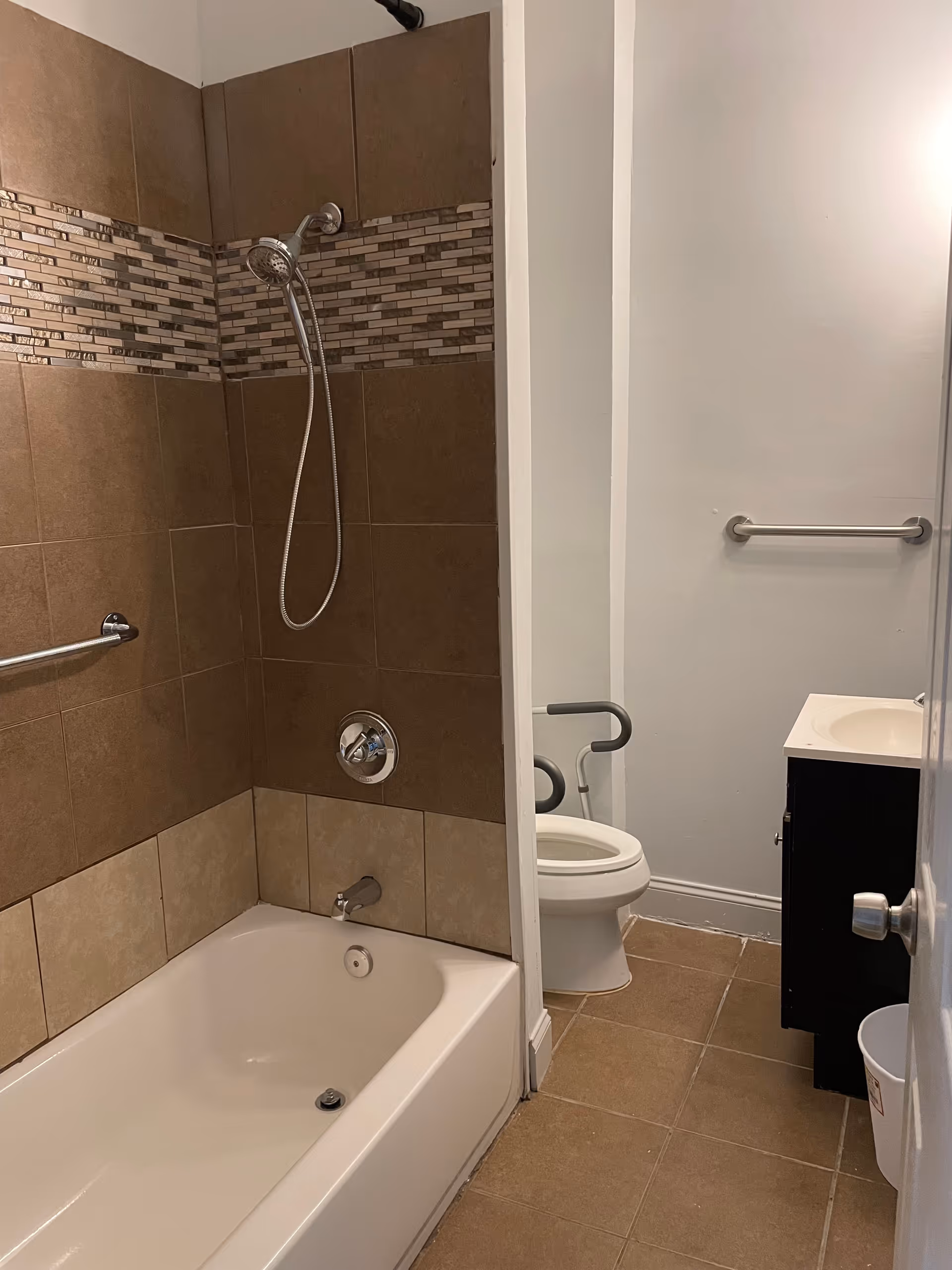 Bathroom with a tiled bathtub and shower, a toilet with grab bars, and a small sink vanity.