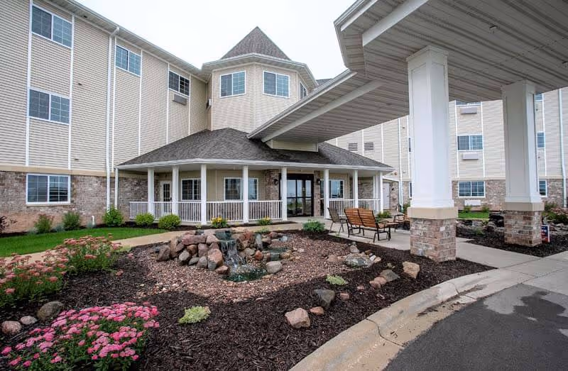 Exterior view of a senior living facility with beige siding and stone accents. The building features a covered entrance supported by white columns with brick bases. In front of the entrance, there is a landscaped area with rocks, a small water feature, and flowering plants. Several benches are placed near the entrance walkway.