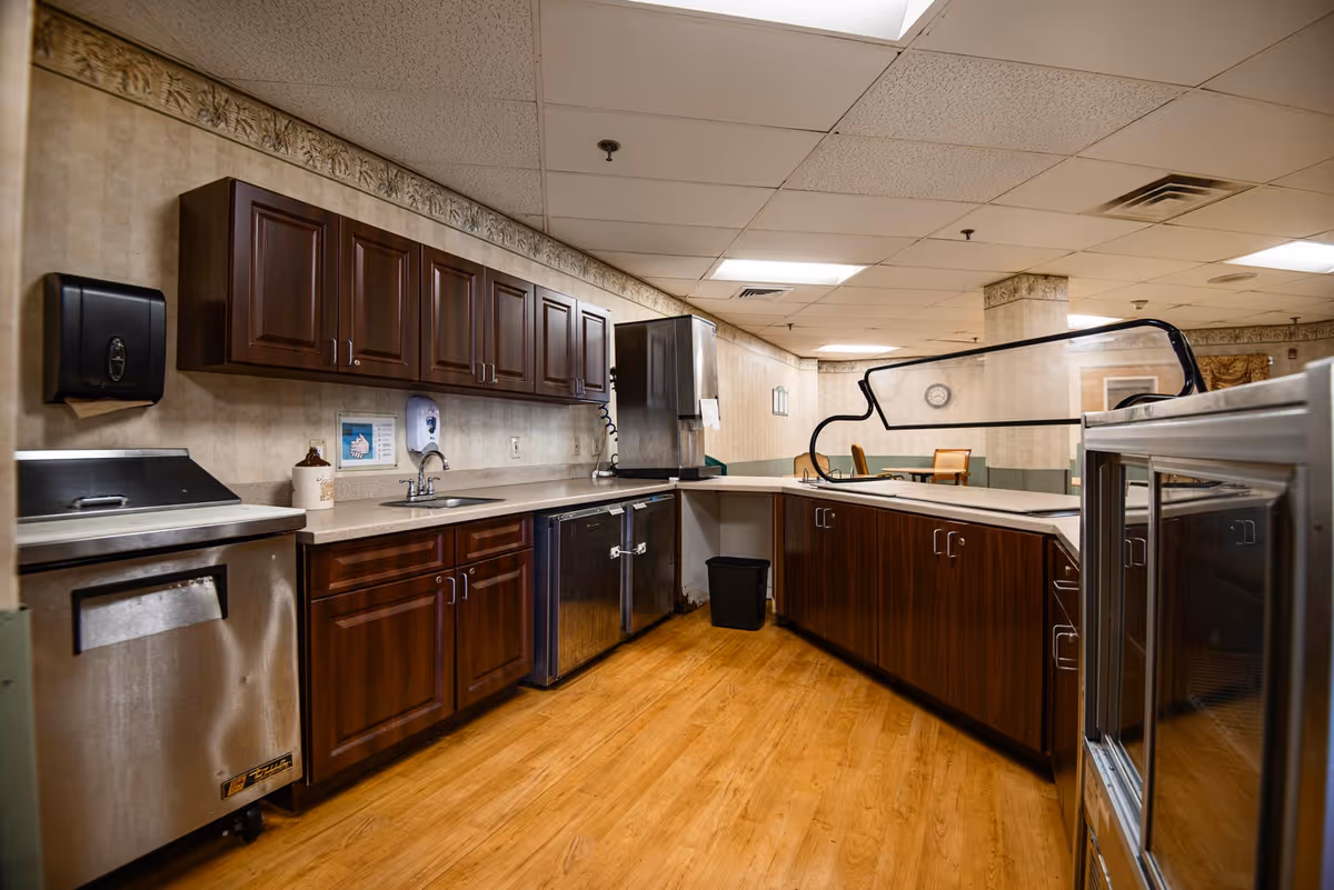 Interior view of a nursing and rehabilitation center kitchen area with wooden cabinets, stainless steel appliances, a sink, and a countertop with a protective glass barrier. The floor is wooden, and the ceiling has fluorescent lighting. In the background, there is a seating area with chairs and a clock on the wall.