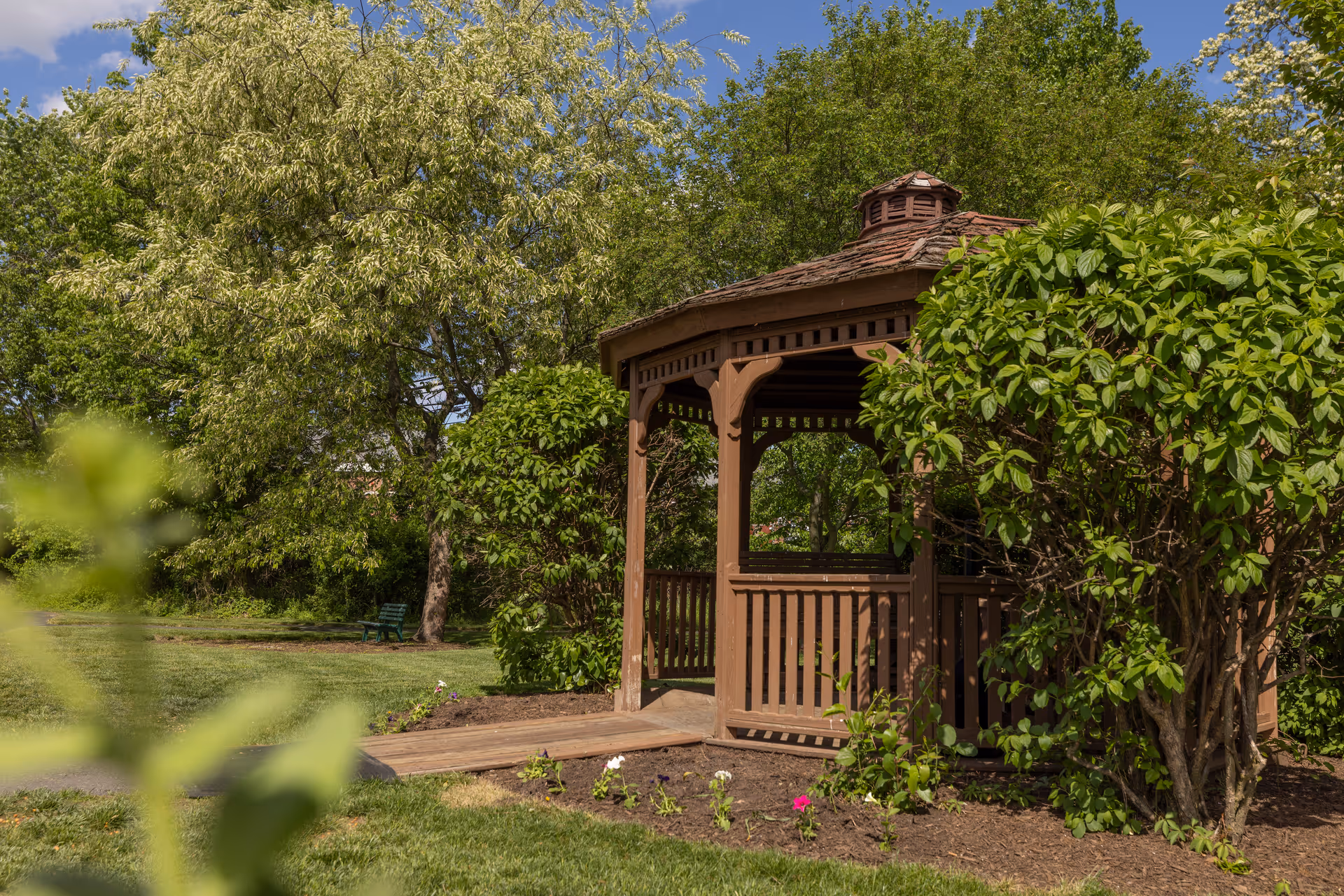 A wooden gazebo surrounded by lush green bushes and trees in a garden under a partly cloudy blue sky.