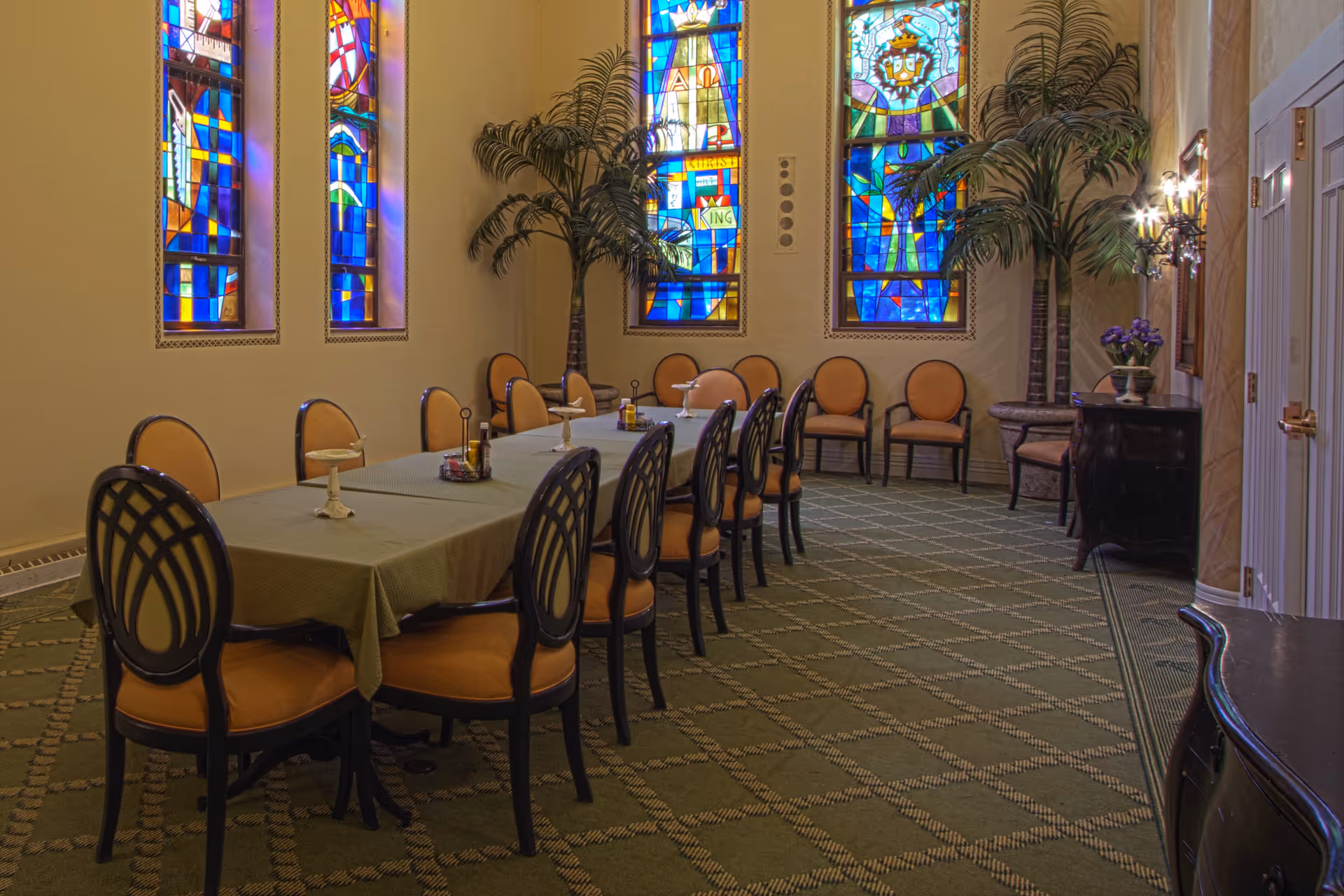 Long dining table with chairs in a formal room featuring stained glass windows and potted palms.