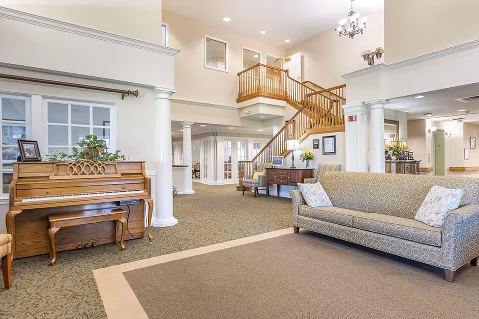A spacious and well-lit senior living facility common area featuring a wooden upright piano with a bench, a patterned sofa with floral pillows, a wooden side table with a lamp and flowers, and a staircase with wooden railings leading to an upper floor. The area has beige walls, carpeted floors, white columns, and a chandelier hanging from the ceiling.