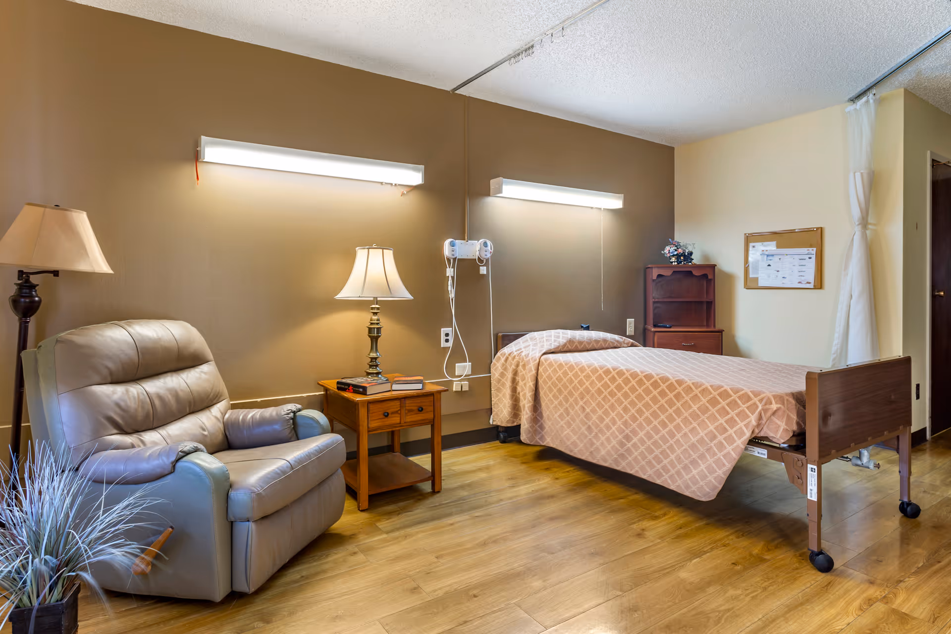 A senior living bedroom with a hospital-style bed, bedside table and lamp, and a recliner chair.