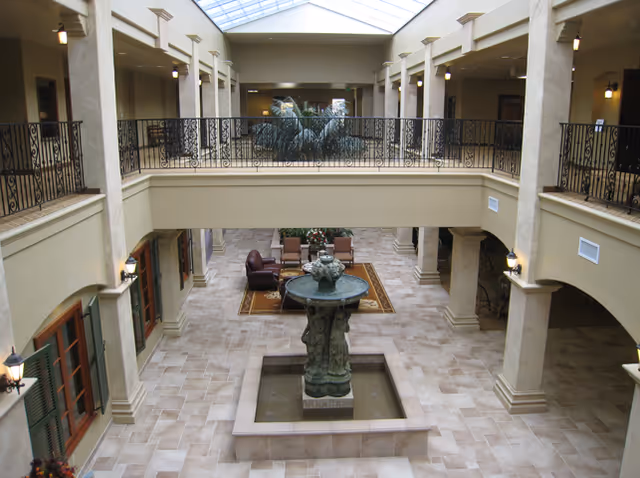 Interior view of a spacious senior living facility atrium with a central decorative fountain, tiled floors, beige walls, and a second-floor balcony with wrought iron railings. There are seating areas with chairs and a table near the far end, and the ceiling features a large skylight allowing natural light to illuminate the space.