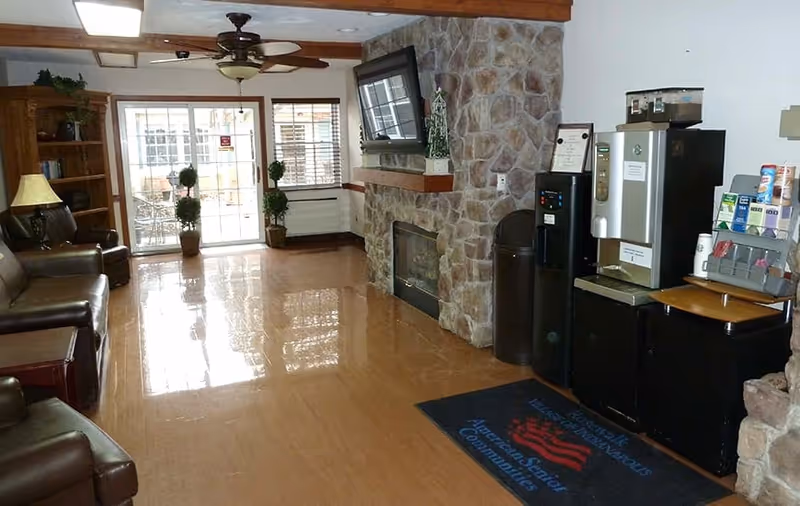 A senior living facility common area with leather chairs, a stone fireplace with a mounted TV above it, a ceiling fan, and a beverage station with coffee and supplies. There are large windows and a glass door leading to an outdoor patio with potted plants.
