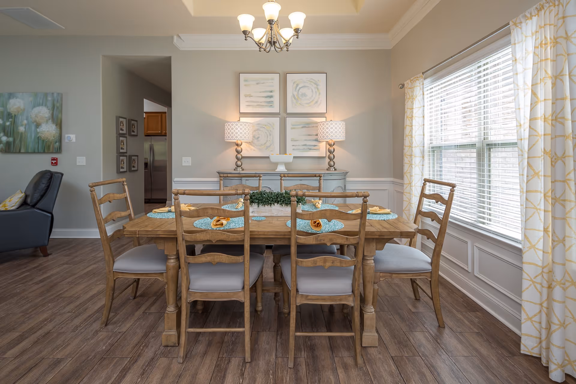 A dining room with a wooden table set for six with blue placemats and yellow napkins. The room has wooden flooring, a large window with white and yellow patterned curtains, a sideboard with two lamps and abstract artwork above it, and a chandelier hanging from the ceiling.