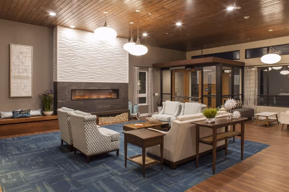 A spacious and modern living room area with a wooden ceiling, recessed lighting, and hanging pendant lights. The room features a contemporary fireplace with a textured white wall above it, surrounded by dark tiles. There are several upholstered chairs and sofas arranged around a wooden coffee table on a blue patterned rug. A wooden console table with decorative items and plants is placed behind one of the sofas. Large windows with curtains allow natural light into the room, and a glass-enclosed entryway is visible in the background.