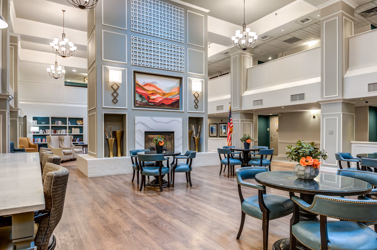 Spacious and well-lit common area in a senior living facility featuring multiple round tables with blue cushioned chairs, a fireplace with decorative vases on either side, a colorful abstract painting above the fireplace, and bookshelves with seating in the background. The room has high ceilings with chandeliers and light wood flooring.