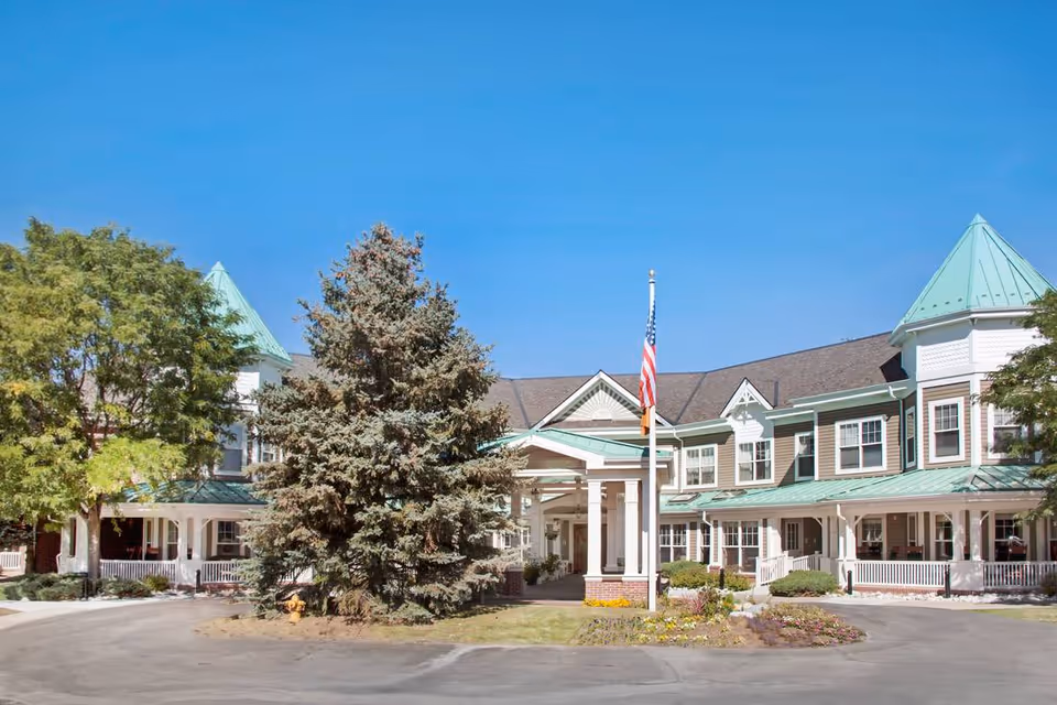 Front exterior view of Sunrise at Pinehurst facility with a circular driveway, large trees, an American flag on a flagpole, and a building with green roofs and white trim under a clear blue sky.