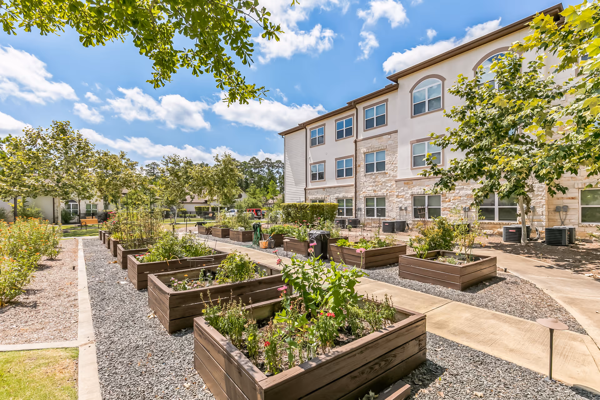 Outdoor garden area at Woodhaven Village featuring multiple raised wooden garden beds with various plants and flowers. The garden is surrounded by paved walkways and trees, with a three-story building in the background under a partly cloudy blue sky.