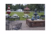 Outdoor seating area with metal chairs and tables, a brick water fountain with decorative pots, picnic tables with umbrellas, a gazebo in the background, and green grass with trees surrounding the space.