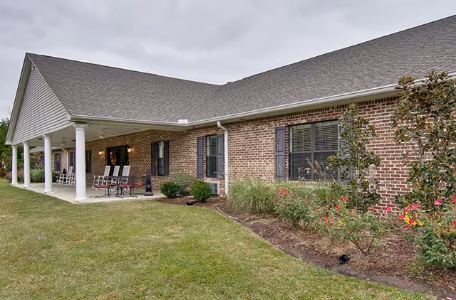 Exterior view of a single-story brick building with a covered porch supported by white columns. The porch has several chairs and a table, and there are bushes and flowers planted along the building's foundation. The sky is overcast.