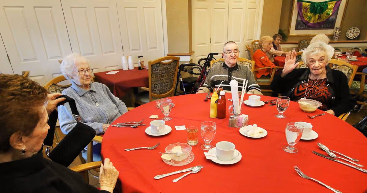 Four elderly residents sit around a round dining table with a red tablecloth in a communal dining room; one woman waves at the camera.