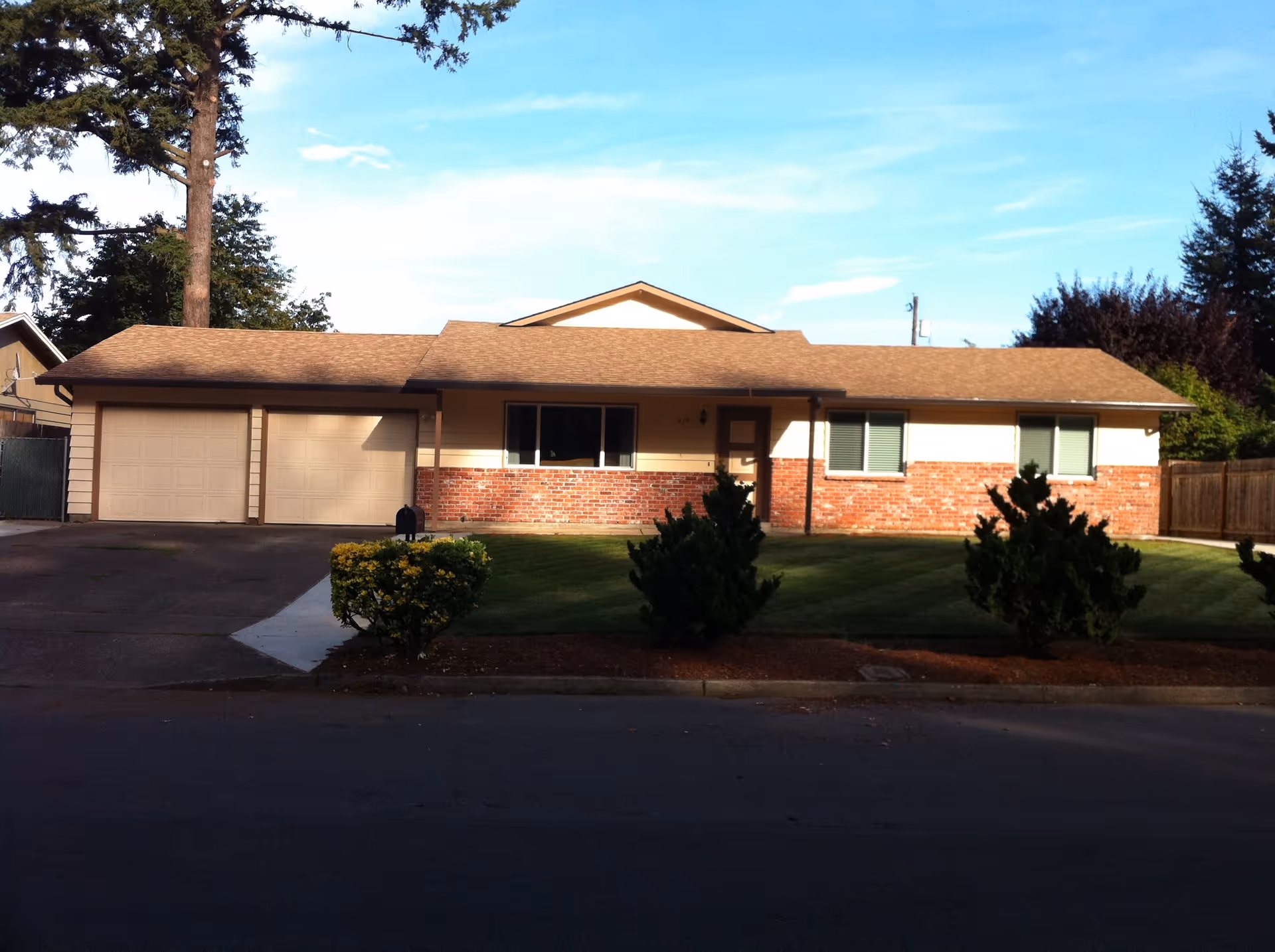 Single-story residential building with a brick and beige exterior, a two-car garage, a front lawn with shrubs, and a clear blue sky above.