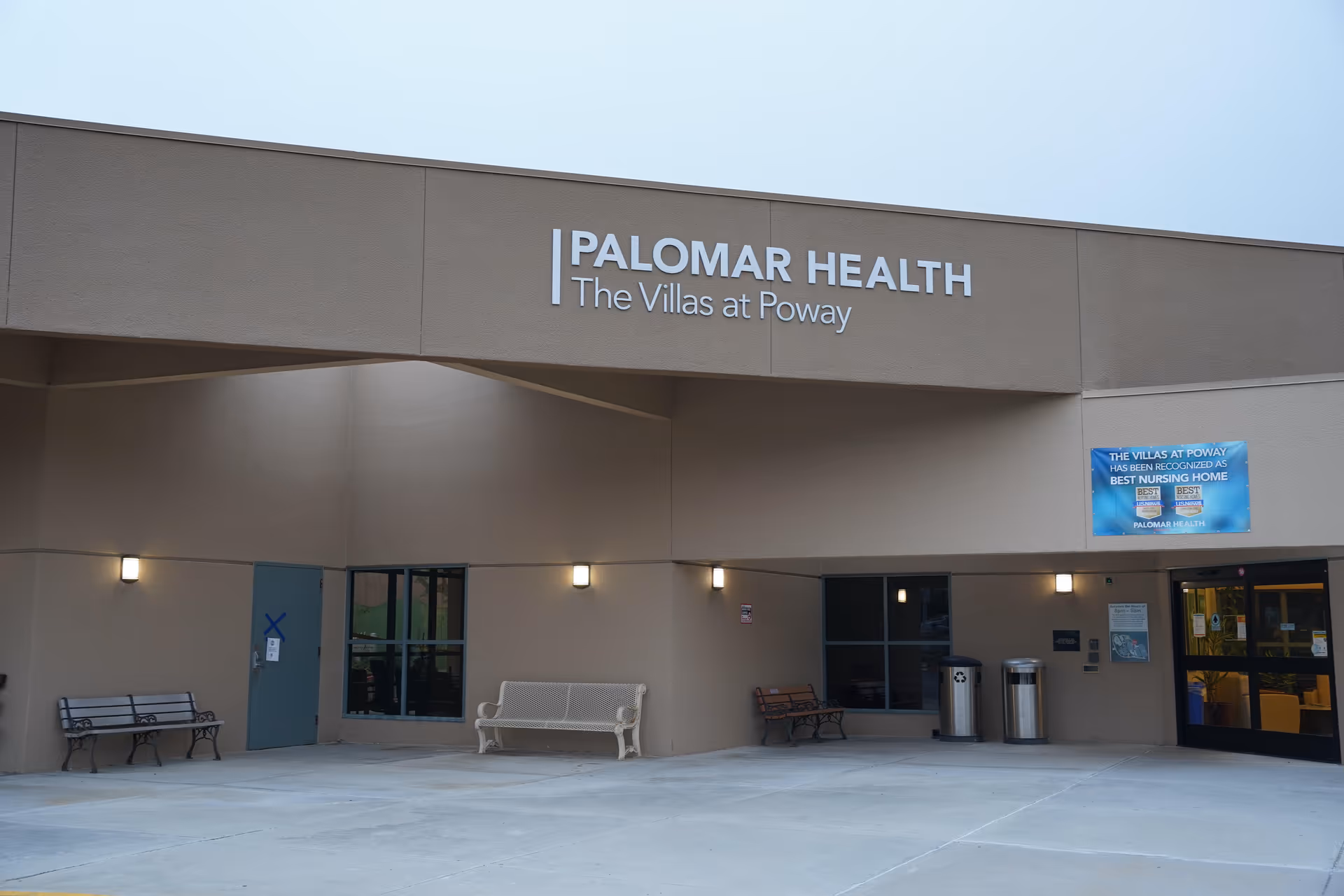 Exterior entrance of Palomar Health The Villas at Poway building with benches, trash bins, and a sign recognizing it as the best nursing home.