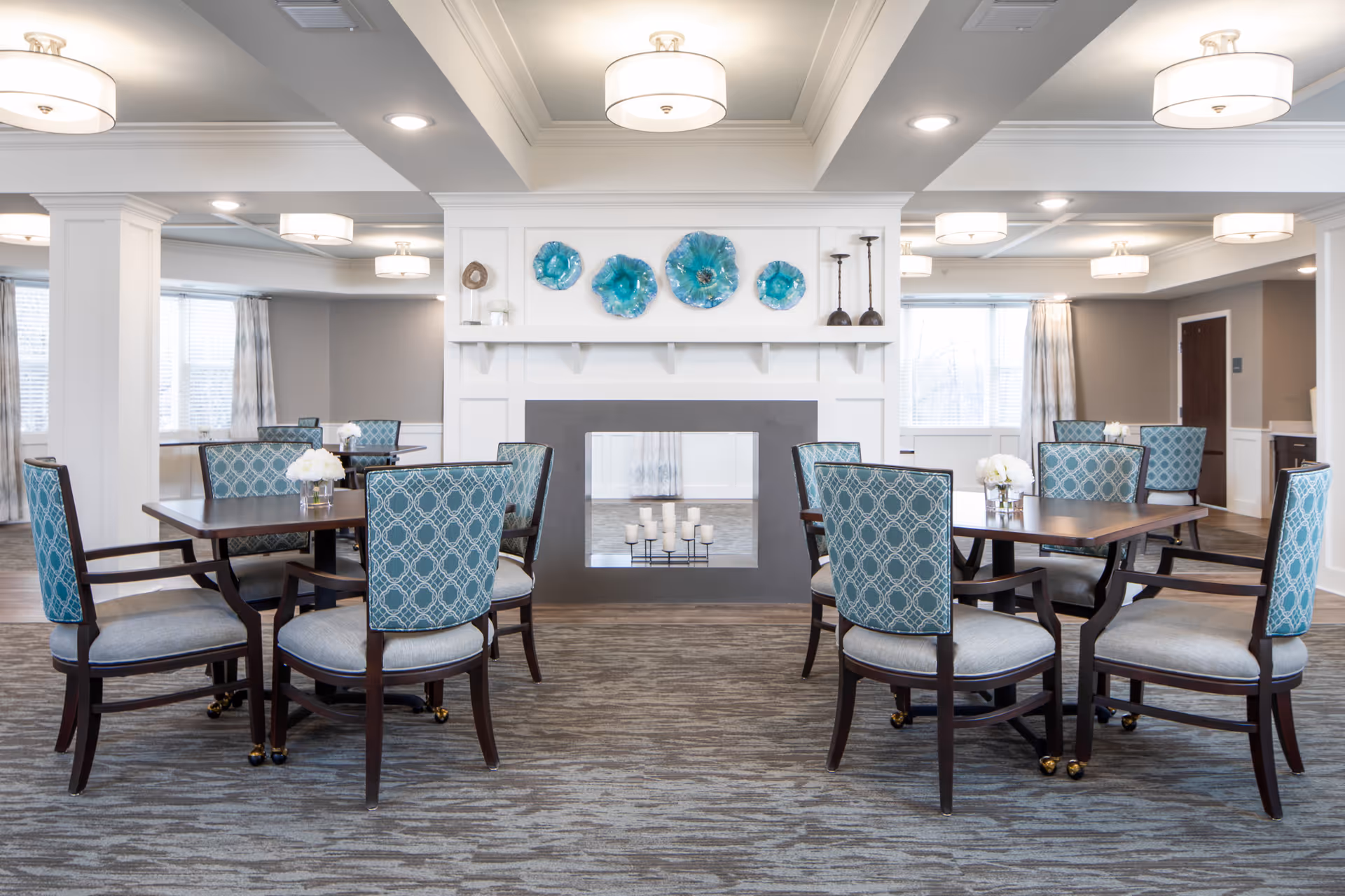 A bright and spacious dining room with multiple wooden tables and upholstered chairs featuring a blue patterned fabric. The room has a modern fireplace with decorative blue glass art pieces mounted above it. Large windows with curtains allow natural light to fill the space, and ceiling lights provide additional illumination.