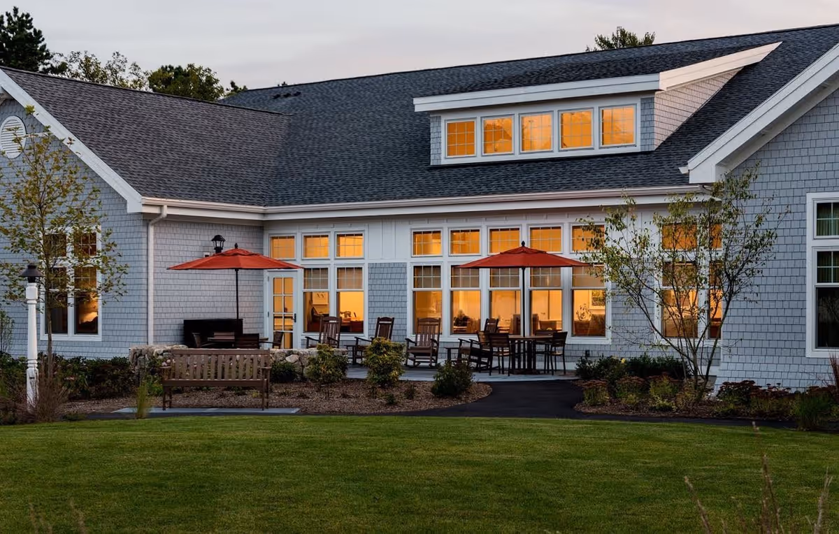 Exterior view of a single-story senior living facility with large lit windows, outdoor seating and red patio umbrellas in a landscaped lawn at dusk.