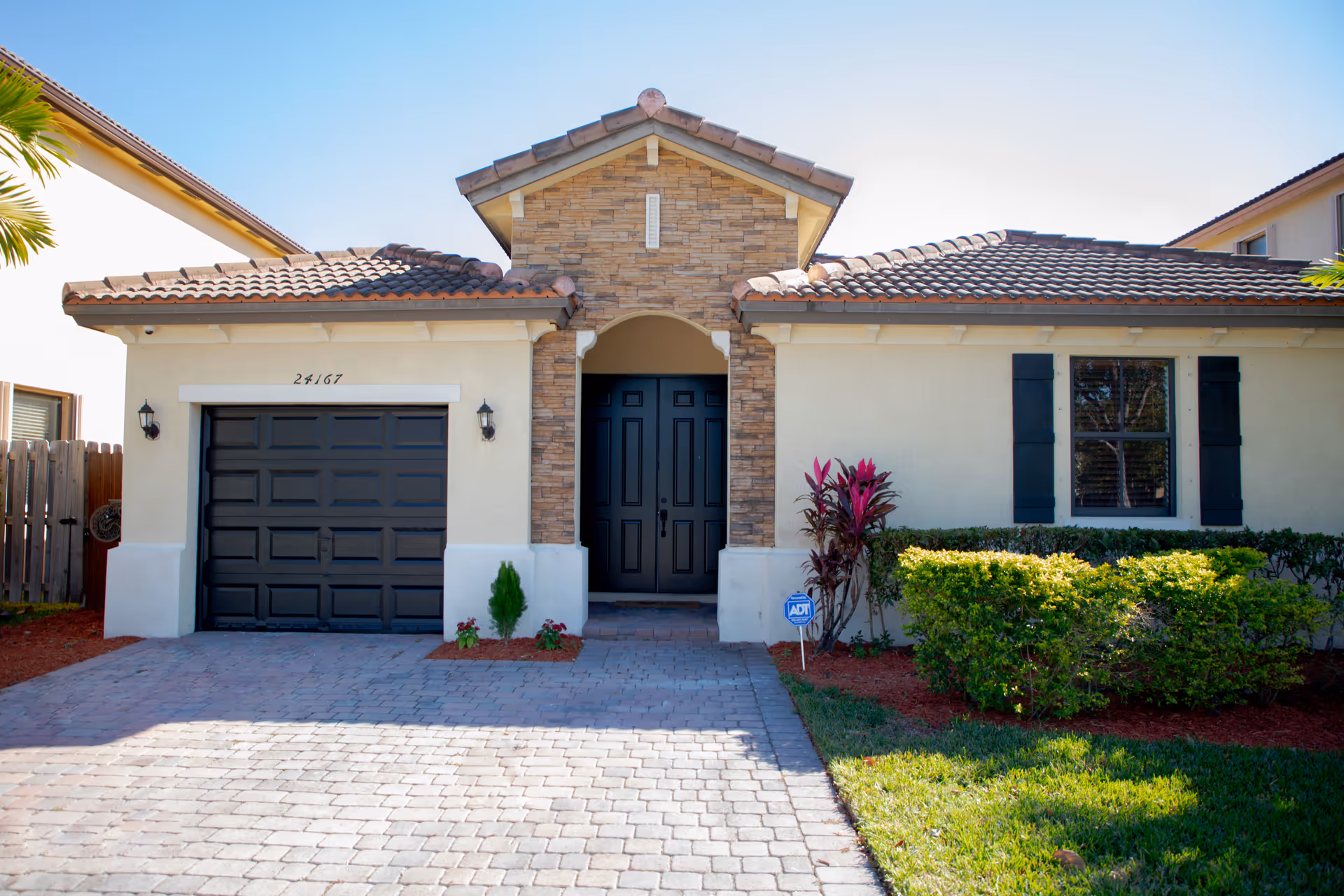 Front exterior view of a single-story house with a tiled roof, a black garage door on the left, a black front door in the center with stone accents around it, and a window with black shutters on the right. The driveway is paved with bricks, and there are small plants and bushes along the front of the house.
