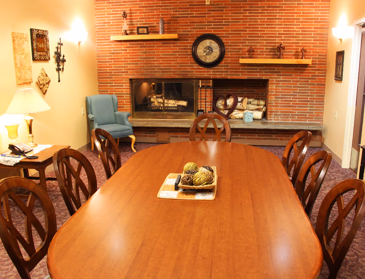 Long wooden dining table with chairs in a cozy common room facing a brick fireplace and wall clock.