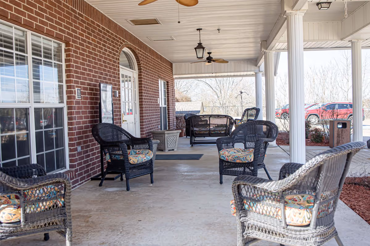 Covered outdoor patio area with wicker chairs and cushioned seats arranged for seating. The patio has a brick wall with windows and a door, white columns supporting the roof, ceiling fans, and a view of parked cars and trees in the background.