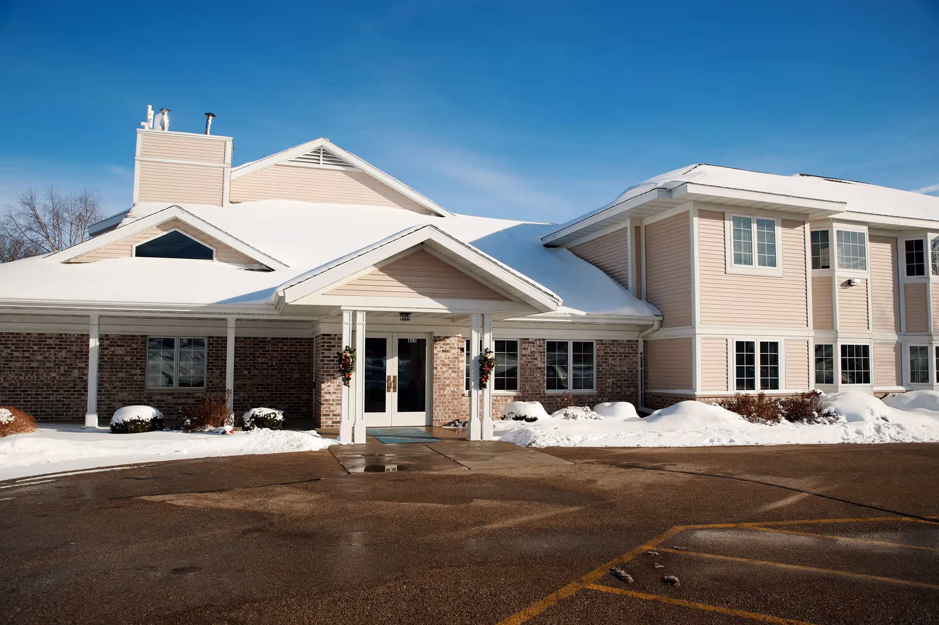 Exterior view of Ingleside Manor, a two-story building with beige siding and brick accents, covered with snow on the roof and surrounding ground under a clear blue sky.