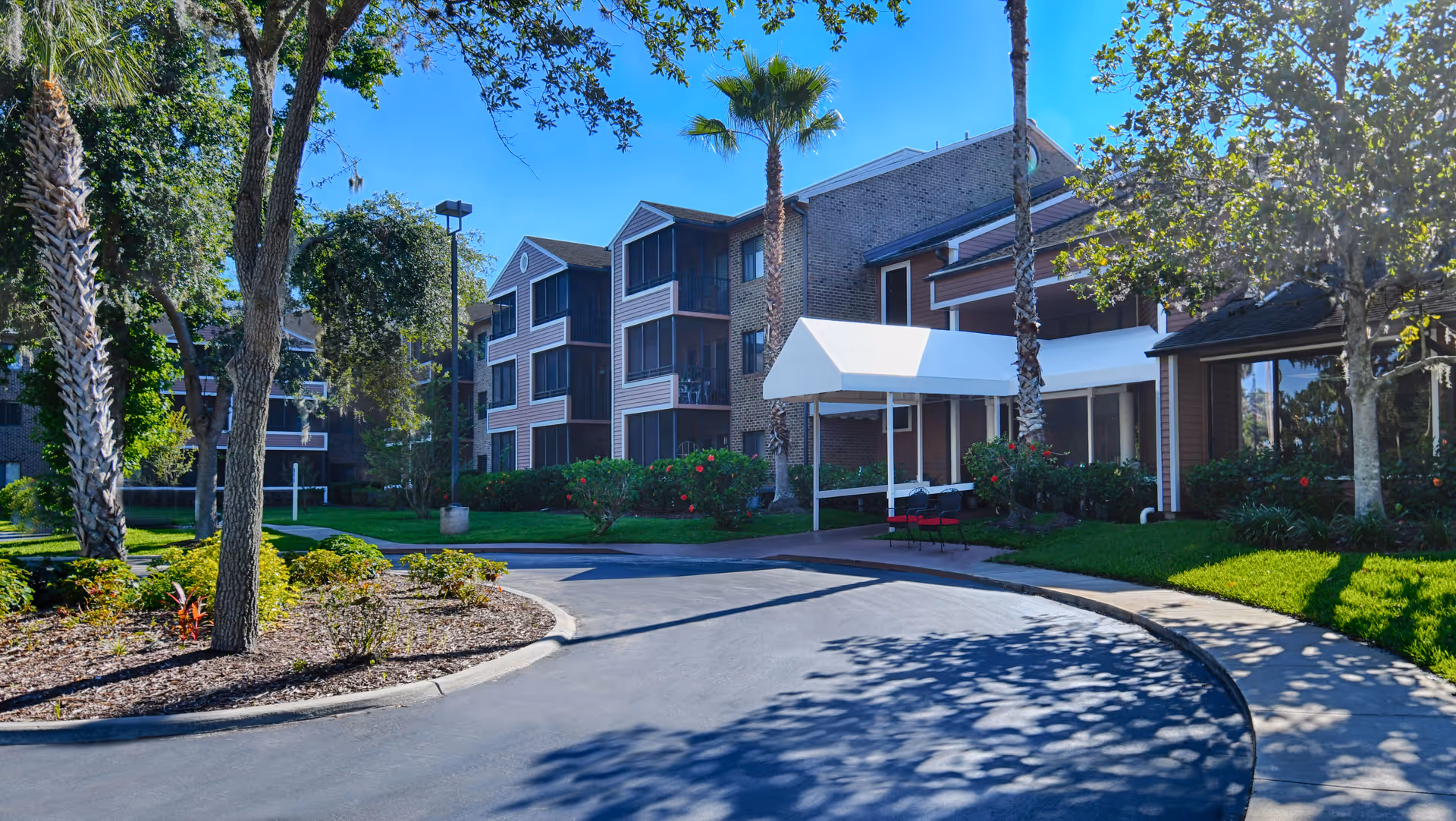 Exterior view of a senior living facility named CountrySide Lakes showing a curved driveway leading to the entrance with a white canopy. The building is three stories tall with screened balconies, surrounded by palm trees, green lawns, and landscaped garden beds under a clear blue sky.