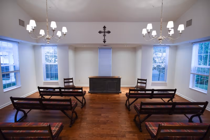 A small chapel or meditation room with wooden benches arranged in rows facing a wooden altar. The room has hardwood floors, white walls, three tall windows with blinds, two chandeliers with lit lamps, and a decorative cross mounted on the wall above the altar.