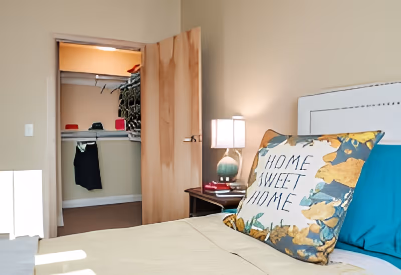 A cozy bedroom with a bed featuring a decorative pillow that says 'Home Sweet Home'. Next to the bed is a nightstand with a lamp and some books. The door to a walk-in closet is open, showing clothes and storage inside.