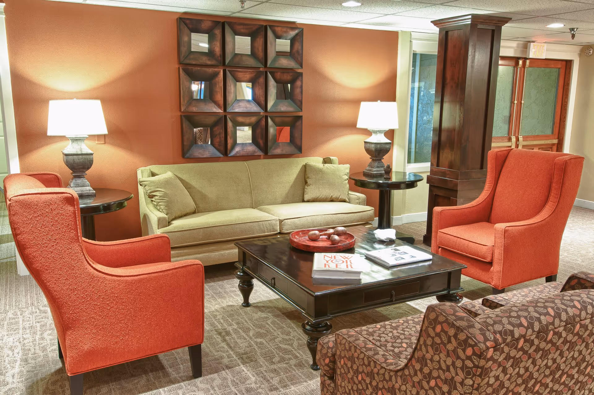 A cozy living room area with a beige sofa, two red armchairs, and a patterned armchair surrounding a dark wooden coffee table. The coffee table has books and decorative items on it. Two table lamps with white shades sit on round side tables next to the sofa. The walls are painted a warm orange color, and there is a decorative wall art piece above the sofa. The room has carpeted flooring and a wooden pillar near the entrance door.