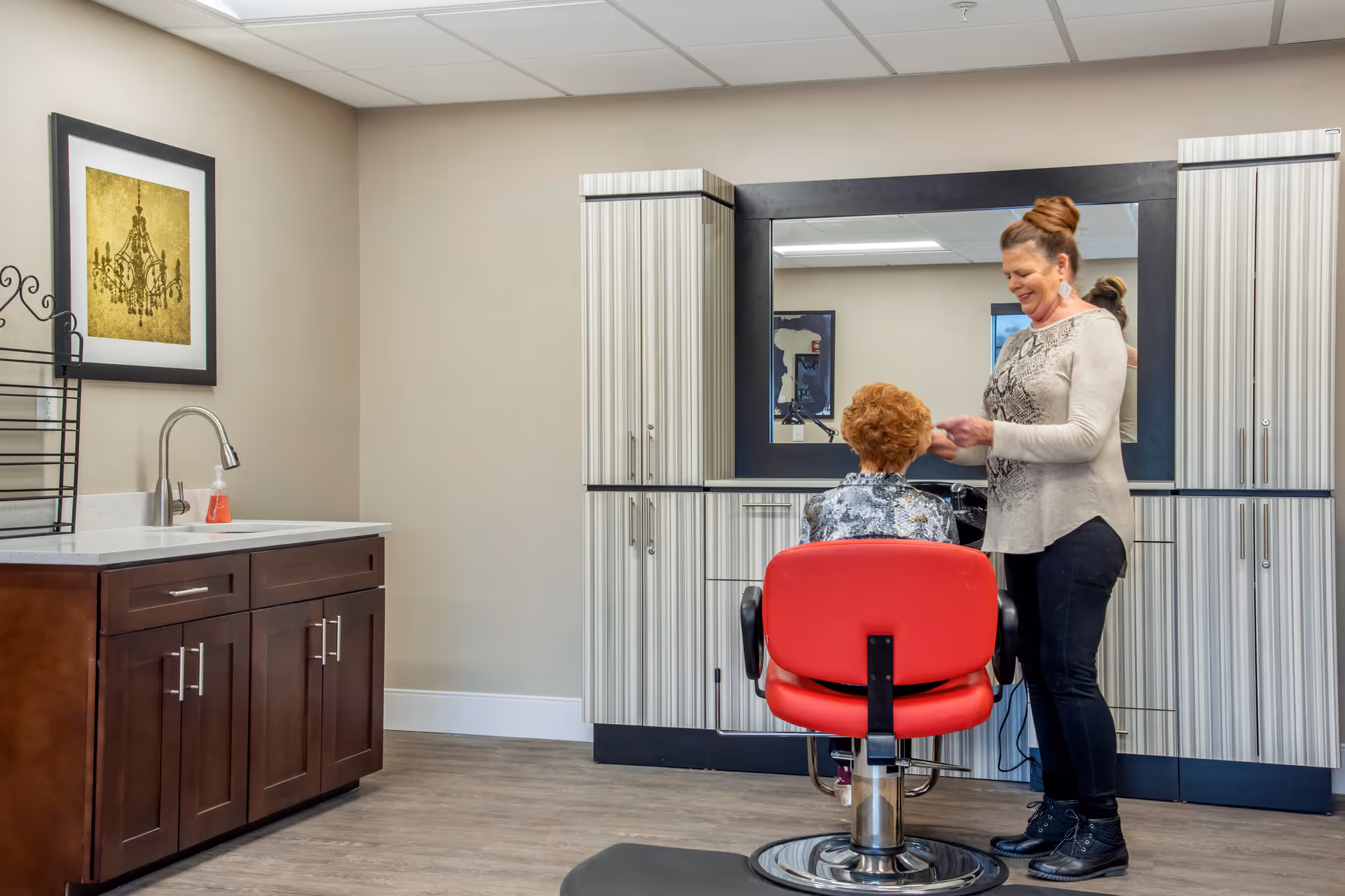 A senior woman sitting in a red salon chair facing a large mirror while another woman stands beside her, styling her hair in a well-lit room with wooden cabinets and a sink on the left side.