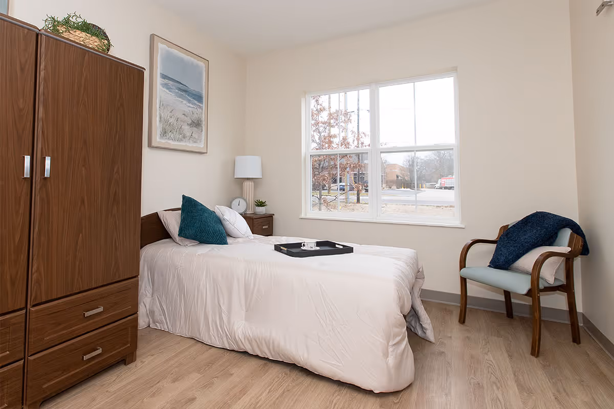 A bright, tidy bedroom with a single bed, wooden wardrobe, nightstand, armchair and a large window.