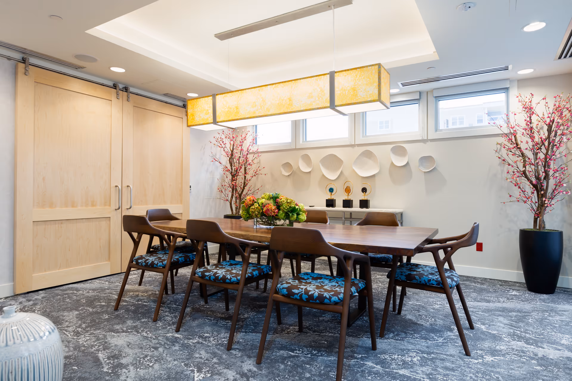 A modern dining room with a wooden table and patterned chairs, a rectangular overhead light, wall decor and potted flowering plants.